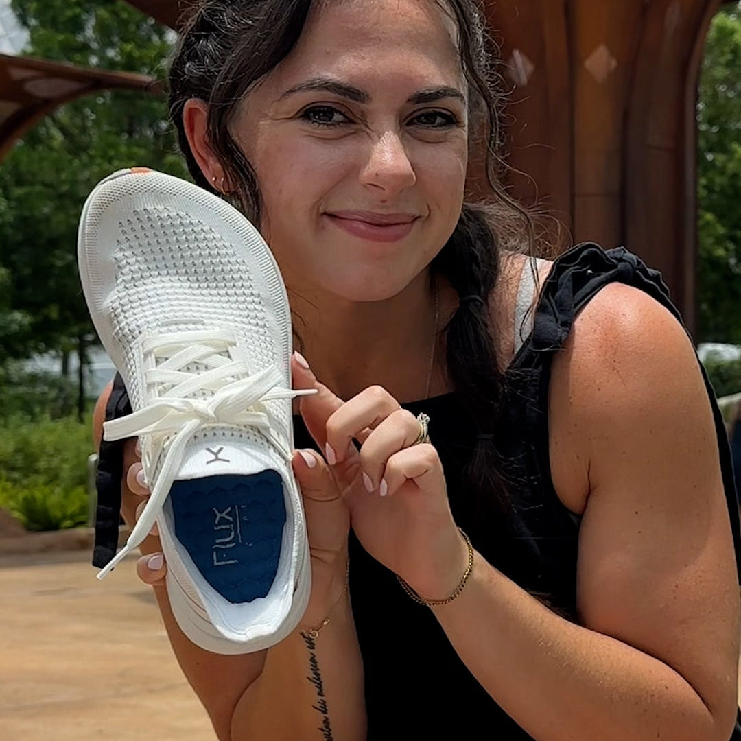 Woman holding a white sneaker with a Flux brand logo outdoors