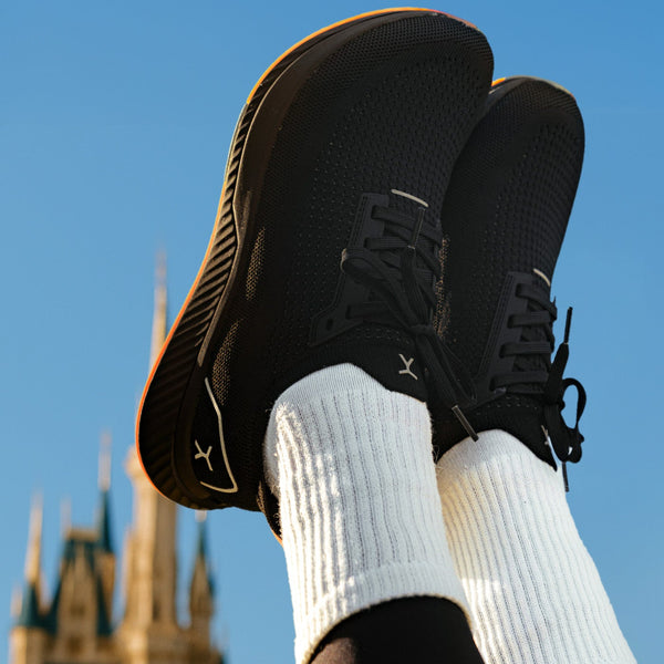 Black sneakers with white socks held up against a blue sky with a castle-like structure in the background